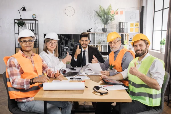 Nitelikli mimarlar, mühendisler ve tasarımcılar TV ekranının önünde iş toplantısı yapıyorlar. Takım elbiseli ve miğferli çok kültürlü meslektaşlar masada oturup kameraya bakıyorlar..