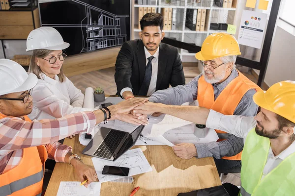 Happy multiracial architects, engineers and designers stacking their hands together during successful meeting at boardroom. Men and women colleagues leading meeting for architectural project.