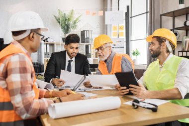 Çok ırklı adamlar resmi kıyafetler ve miğferlerle önemli stratejik oturumlar sırasında bir sürü planla masada oturuyorlar. Ortak inşaat projesinde çalışan iş adamları.