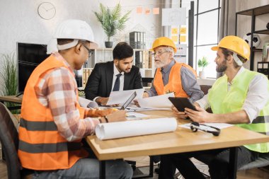 Çok ırklı adamlar resmi kıyafetler ve miğferlerle önemli stratejik oturumlar sırasında bir sürü planla masada oturuyorlar. Ortak inşaat projesinde çalışan iş adamları.