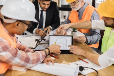 Ofisteki iş toplantısı sırasında inşaat ekibinin planlarını analiz et. İş için dijital tablet kullanan çok ırklı ortaklar. İşbirliği ve beyin fırtınası kavramı.
