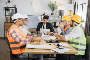 Çeşitli mimarlar, tasarımcılar ve mühendisler büyük bir plazma TV ekranının önünde modern cihazlar ve planlarla masada oturuyorlar. Ortak inşaat projesi üzerinde çalışan iş adamları.
