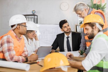 Koruyucu şapkaya odaklan. İş yerindeki konferansı yöneten uzman mühendisler, tasarımcılar ve mimarlar. Takım elbiseli, üniformalı ve koruyucu başlıklı çok kültürlü insanlardan oluşan bir grup..