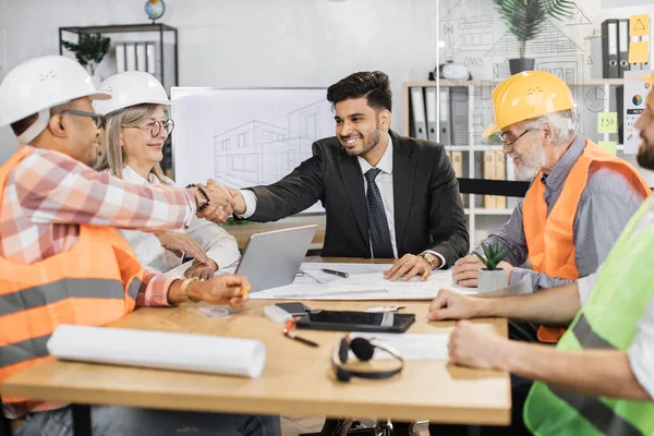 Çok ırklı mimarlar, tasarımcılar ve mühendisler masada oturup el sıkışarak birbirlerini selamlıyorlar. İş ortakları büyük TV plazma ekranıyla ofiste toplantı yapıyorlar.