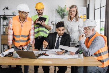 Miğferli çok ırklı mimarlar planlar ve modern cihazlarla masada oturuyorlar. Odaklanmış işçiler yeni bir inşaat projesi oluşturmak için işbirliği yapıyor.