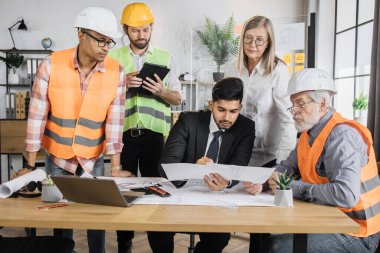 Miğferli çok ırklı mimarlar planlar ve modern cihazlarla masada oturuyorlar. Odaklanmış işçiler yeni bir inşaat projesi oluşturmak için işbirliği yapıyor.