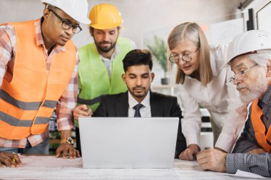 Dört erkek ve bir kadın birlikte masada oturmuş iş konferansı sırasında dizüstü bilgisayarın planlarını tartışıyorlar. Ofiste çalışan çok ırklı mimarlar, tasarımcılar ve mühendisler.