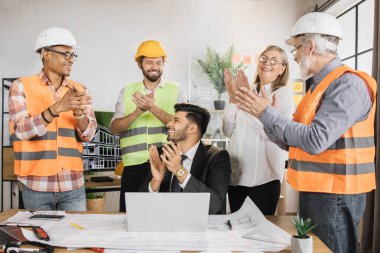Çok uluslu mühendislerden oluşan bir ekip, başarılı bir projeden sonra dizüstü bilgisayara baktıkları için seviniyor. Mühendisleri yanında dururken Hindu patron masada oturuyor..