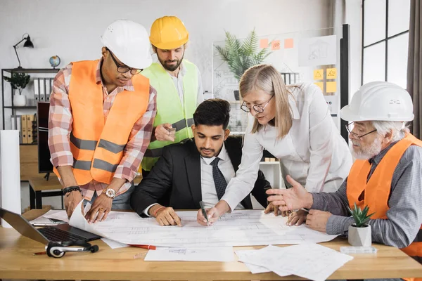 Takım elbiseli Hintli adam yeni inşaat projesini ofisteki kask ve üniformalı meslektaşlarına sunuyor. Masanın yanında duran üç erkek ve bir kadın yetenekli partnerlerini dinliyorlar..