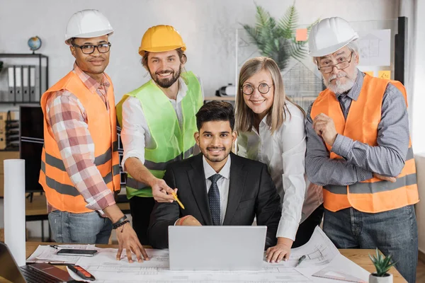 Designers, architects and engineers gathering at office for creating project of new building. Partners of various ages, gender and races standing near or sitting at table and working with blueprints