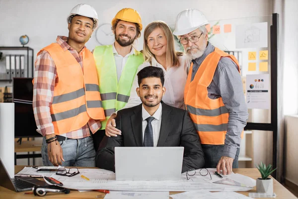 Team of qualified architects, engineers and designers having meeting in boardroom at office indoor, standing near indian man boss, sitting on desk with laptop, looking at camera.