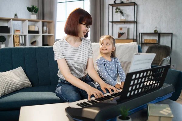 Mom and little happy girl in music therapy by playing piano on music room. Teacher helping young female pupil in piano lesson. Relaxing at home.