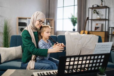 Tesettürlü genç ve güzel Müslüman bir kadın ve sevimli küçük kızı evde flüt çalarken gülümsüyorlar. Annem küçük sevimli müzisyen kıza flüt çalmayı öğretiyor..