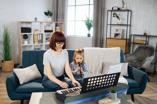 Beautiful young woman playing classic digital piano and her charming little daughter are smiling while playing flute at home. Mother teaching cute little musician girl to playing musical instruments.
