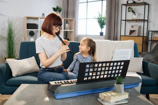 Mom and little happy girl in music therapy by playing flute on music room. Teacher helping young female pupil in flute lesson. Relaxing at home.