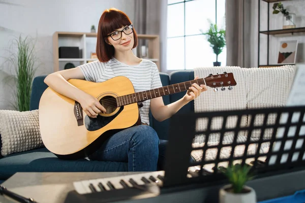 Young woman plays the guitar on bright living room at home. Girl sitting on sofa with a musical instrument indoor. Pretty young woman study to play guitar.