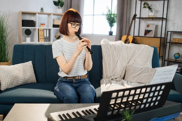 Young woman plays the flute on bright living room at home. Girl sitting on sofa with a musical instrument indoor. Pretty young woman study to play flute.