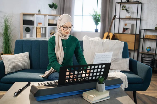 Young muslim woman in hijab plays the piano on bright living room at home. Girl sitting on sofa with a musical instrument indoor. Pretty young arabic woman study to play piano.