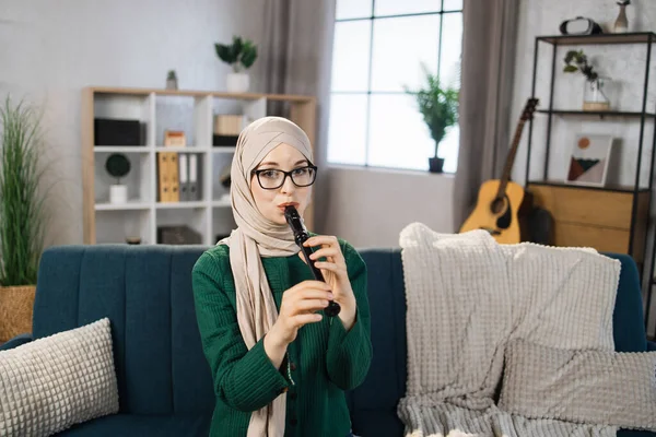 Portrait of young arab female with hijab playing on flute. Pretty muslim girl learning to play flute at home. Beautiful student woman playing musical instrument.