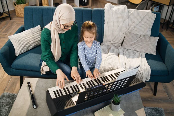 Portrait of muslim woman in hijab and her cute little daughter learning to play piano at home. Teacher teaching pretty girl to play piano in classroom.