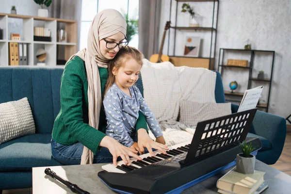 Child girl learning to play piano using notes together with her muslim mother in hijab. Remote learning from home. Arts for kids. Little girl with musical instrument. Creative girl play song.