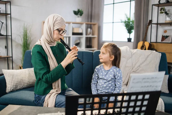 Muslim mom and little happy girl in music therapy by playing flute on music room. Teacher helping young female pupil in flute lesson. Relaxing at home.