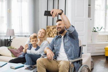 Modern oturma odasında mutlu bir eş ve okul çağındaki oğluyla akıllı telefon kamerasında poz veren erkek tekerlekli sandalye kullanıcısı. Genç bir aile birlikte zaman geçirir ve rahat bir evde selfie çeker..