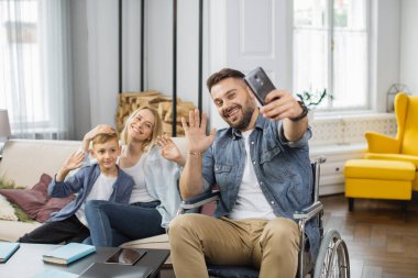 Kafkasyalı engelli bir adam akıllı telefon tutuyor ve ailesiyle selfie çekiyor. Gülümseyen anne ve oğul birlikte koltukta oturuyor, tekerlekli sandalyedeki babanın yanında ve telefon kamerasıyla el sallıyorlar..