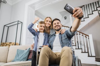Gülümseyen aile, özel ihtiyaçları olan babanın yanında duruyor ve çevrimiçi video görüşmesi sırasında akıllı telefon kamerasına bakıyor. Baba tekerlekli sandalyede telefon tutarken okul çocuğu el sallıyor..