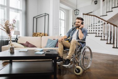 Genç engelli adam evde sevdiği şarkıları dinlemek için kulaklık kullanıyor. Tekerlekli sandalyedeki sakallı erkek hafta sonları boş zamanlarında iyi müzik dinliyor..