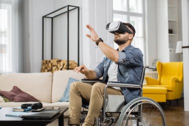Tekerlekli sandalyede otururken ve evde 3 boyutlu oyunlar oynarken gözlük takan beyaz bir adam. Engelli ve modern teknolojiye sahip insanlar için eğlence anlayışı.