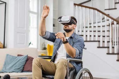 Tekerlekli sandalyedeki sakallı adam sanal gerçeklik kulaklığı kullanarak hayali ekranda hareketler yapıyor. Özel ihtiyaçları olan beyaz erkek evde favori oyunu oynuyor..