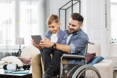Tekerlekli sandalyede oturan engelli bir baba, parlak oturma odasında modern tablette oynarken oğlunu dizlerinin üzerine çöktürüyor. Kafkas aile dostları evde oyun oynuyorlar..
