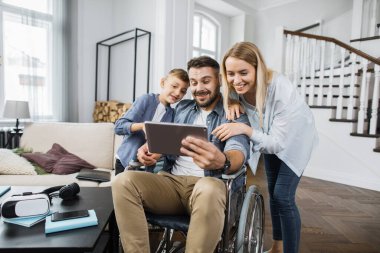 Tekerlekli sandalyede elinde modern tablet olan mutlu bir baba ve yanında oğlu olan eşine komik videolar gösteriyor. Moda oturma odasındaki cihaz ekranına bakarken neşeli aile gülümsüyor.