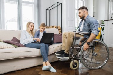 Tekerlekli sandalyedeki engelli adam, sevgi dolu karısı ve güzel okul çağındaki oğluyla evde dijital laptop kullanarak konuşuyor. Genç bir kadın ve çocuk rahat bir koltukta oturuyor..
