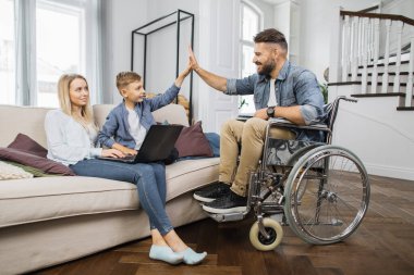 Mutlu beyaz kadın oğluyla birlikte rahat koltukta oturuyor ve modern dizüstü bilgisayar kullanıyor. Tekerlekli sandalyedeki engelli babayla beşlik çakan sevimli çocuk..
