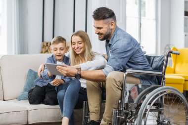 Tekerlekli sandalyedeki beyaz adam ve sevimli oğlu evdeki dijital tablette video izleyen sevimli karısı. Aile kavramı, engellilik ve teknoloji.
