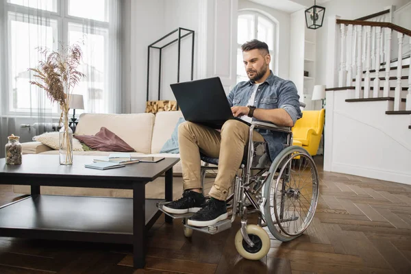 Young male wheelchair user in casual clothes looking at laptop screen during online work in modern living room. Concept of technology and remote job for people with disabilities.
