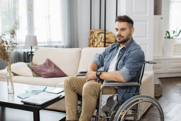 Disabled young man wearing casual clothes sitting in wheelchair at cozy home. Young male getting lost in thoughts during leisure time in living room.