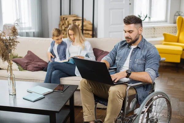 Bearded young man in wheelchair working on laptop while beautiful wife with cute son sitting on couch and reading book. Disabled male working remotely at home with family on background.