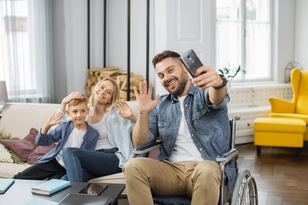 Caucasian man with disabilities holding smartphone and taking selfie with family at home. Smiling mother and son sitting together on sofa near father in wheelchair and waving hands on phone camera.