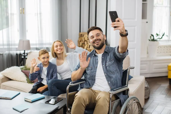 Caucasian man with disabilities holding smartphone and taking selfie with family at home. Smiling mother and son sitting together on sofa near father in wheelchair and waving hands on phone camera.