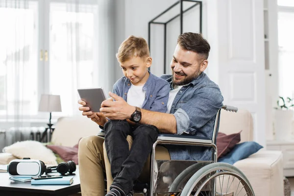 Disabled dad sitting in wheelchair and holding son on knees while playing on modern tablet in bright living room. Friendly caucasian family gaming together at home.