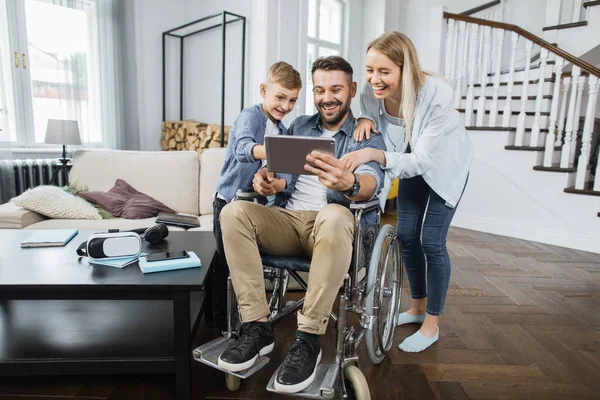 Tekerlekli sandalyede elinde modern tablet olan mutlu bir baba ve yanında oğlu olan eşine komik videolar gösteriyor. Moda oturma odasındaki cihaz ekranına bakarken neşeli aile gülümsüyor.