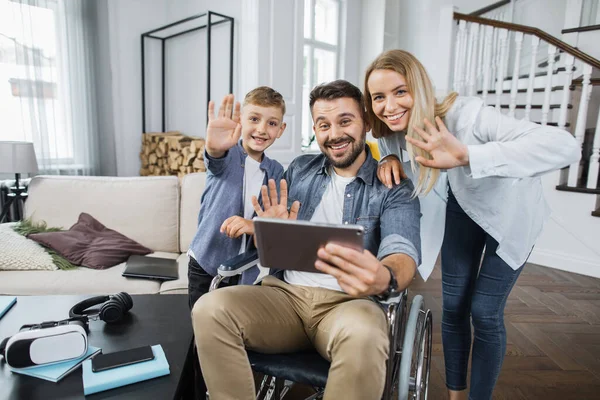 Smiling wife and son standing near disabled father with digital tablet and waving hello while looking at camera. Concept of friendly family and modern leisure entertainment.