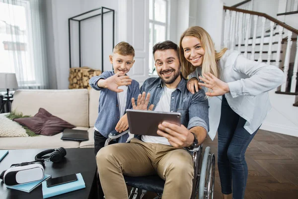 Portrait of disabled husband with blond wife and cute son posing together at cozy apartment. Smiling family waving hello gesture to friends during video call on digital tablet.