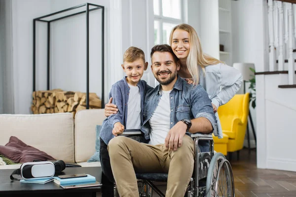 Smiling disabled man in wheelchair posing on camera together with cute son and pretty happy wife in apartment with modern interior. Concept of happy family and support.
