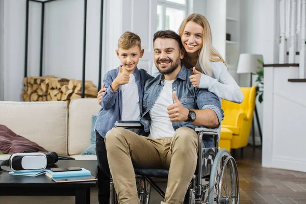 Smiling disabled man in wheelchair posing on camera together with cute son and pretty happy wife in apartment with modern interior. Concept of happy family and support.
