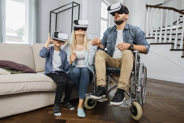 Happy family using modern futuristic technology in bright living room. Caucasian father in wheelchair teaching young wife and cute son how to use virtual reality headset.