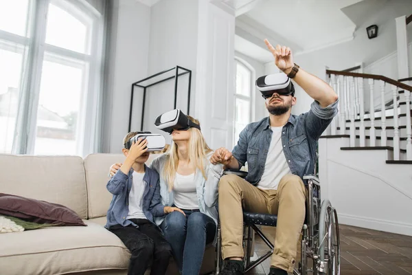 Happy family using modern futuristic technology in bright living room. Caucasian father in wheelchair teaching young wife and cute son how to use virtual reality headset.
